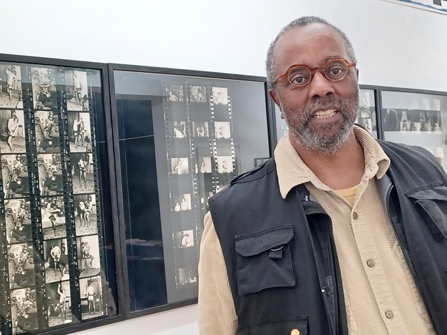 Dr. Reid stands in front of a wall of framed cinema film negatives. He is wearing a tan dress shirt, black vest, and brown-rimmed glasses, and he is smiling at the camera.