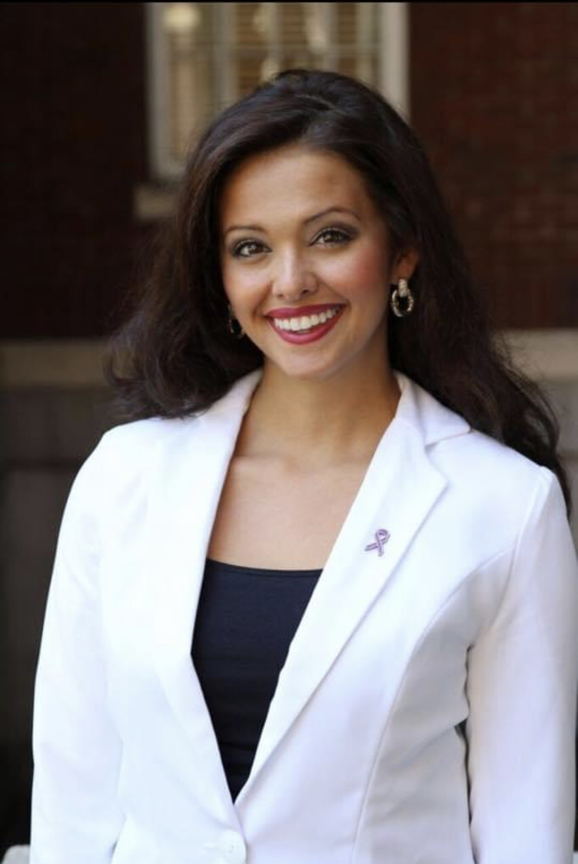 A headshot of Chandler Mordecai. She wears and black blouse with a white blazer, and is smiling broadly.