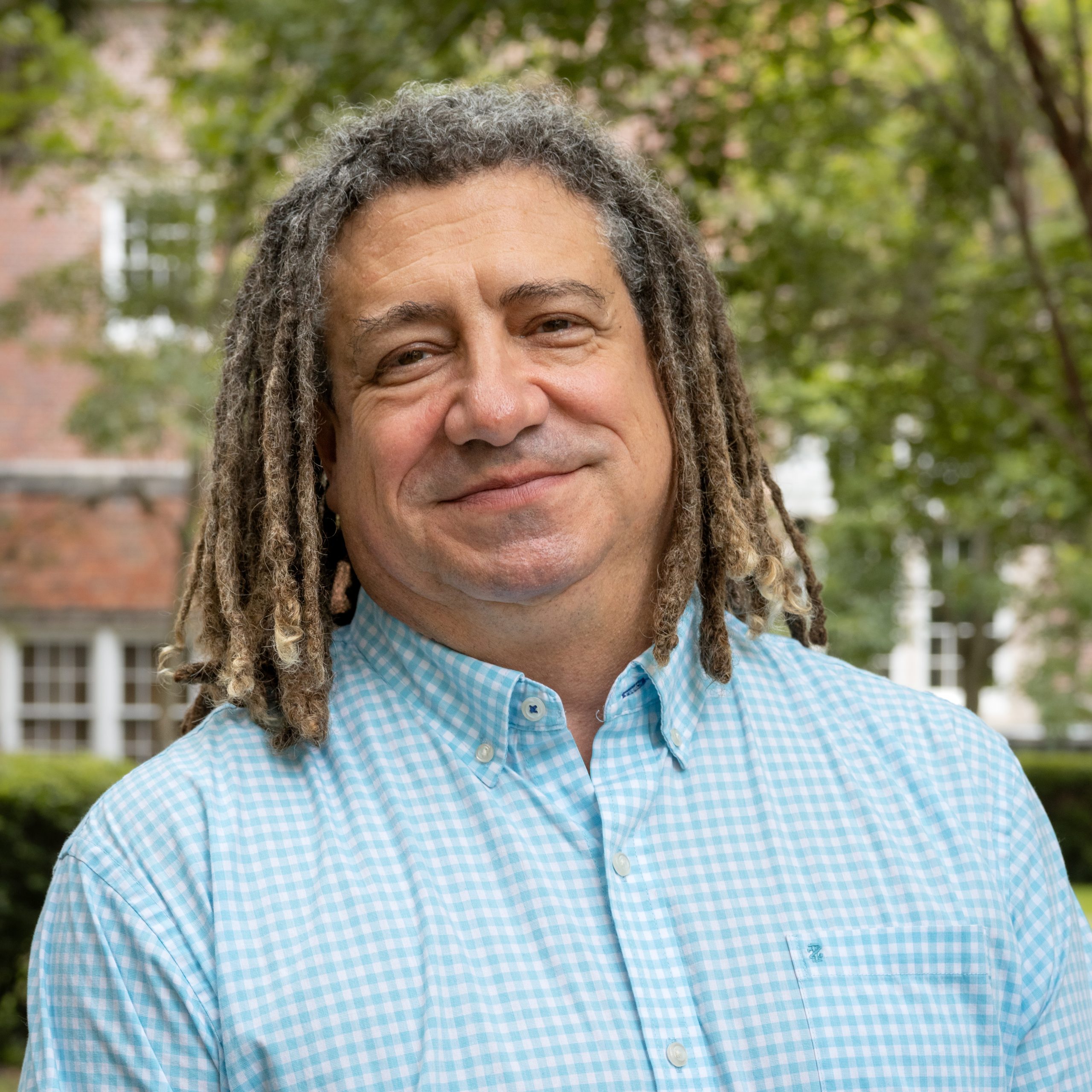 A headshot of Sid Dobrin, who smiles at the camera. He has shoulder-length dreadlocks, and he is wearing a light blue, gingham dress shirt.