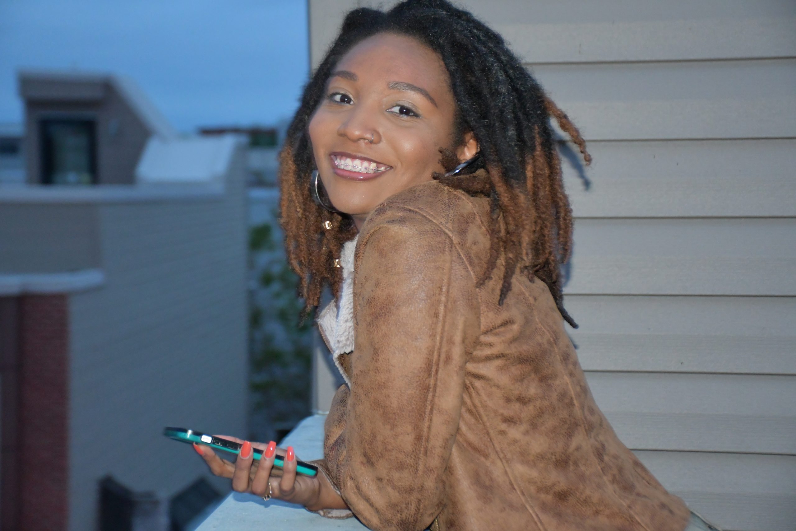 Tiffany smiles while facing the camera. She is holding a cell phone with a green case and has brightly painted orange nails.