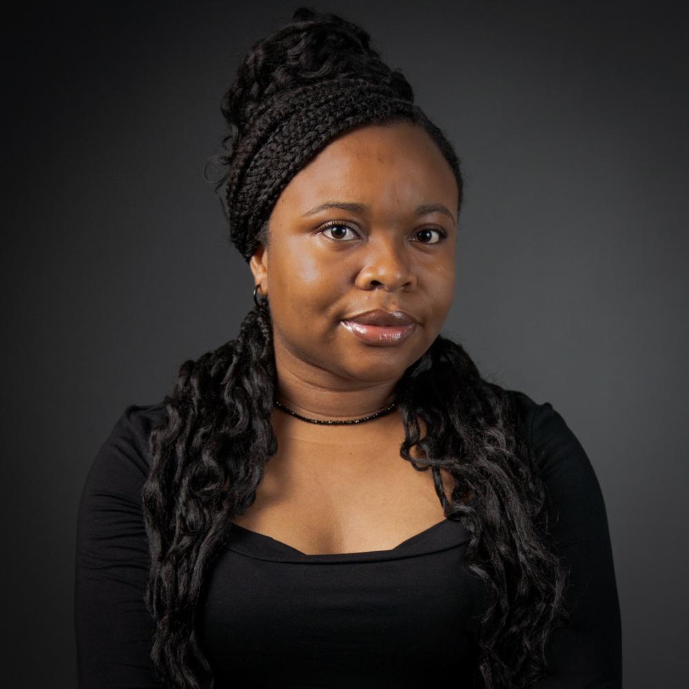 A headshot of Toluwani Odedeyi smiling at the camera. She is a Black woman pictured with her long dark hair in a half updo of braids and curls. She wears a black blouse, simple necklace, and small hoop earrings.
