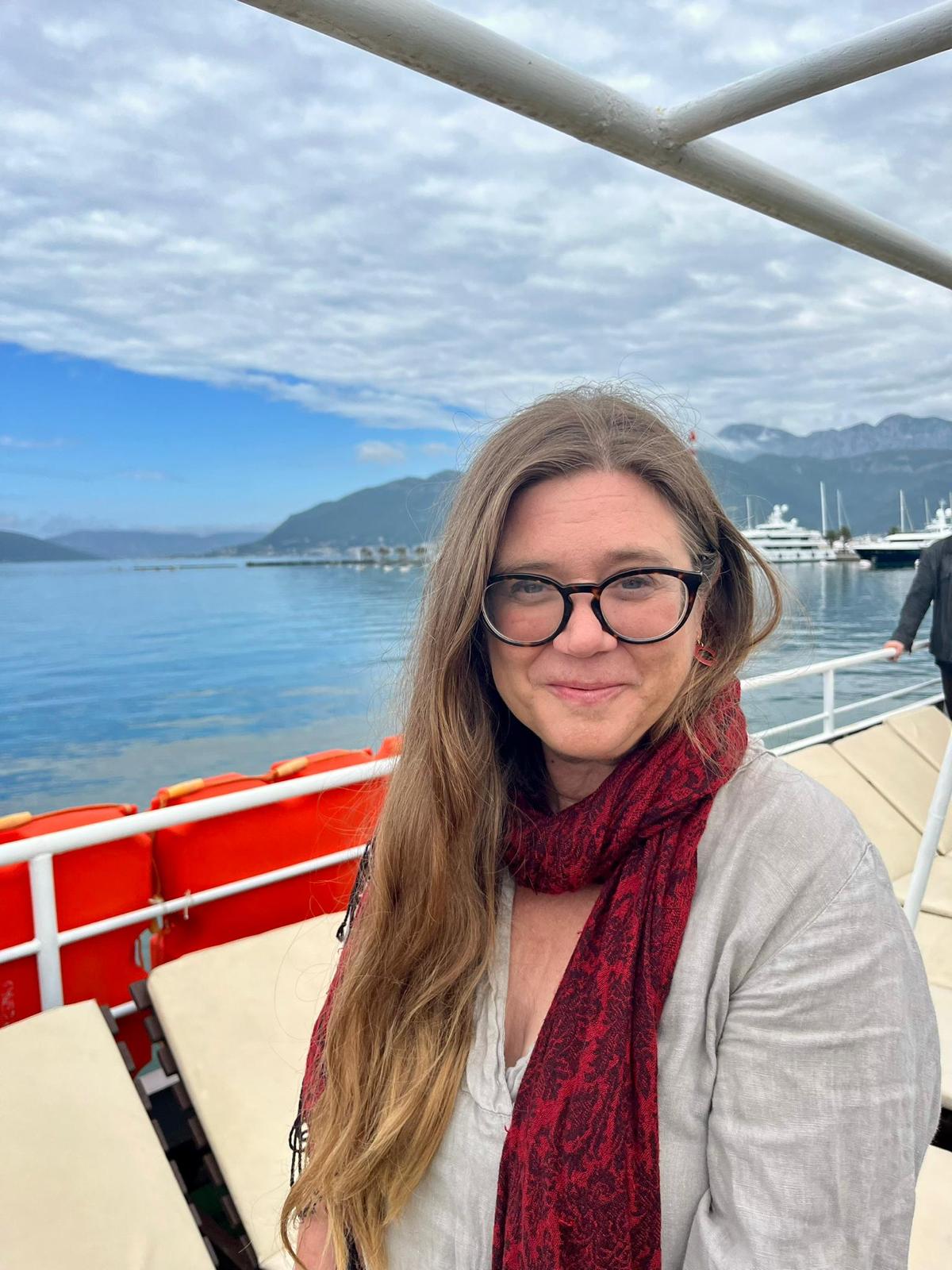 Anastasia Ulanowicz is on a boat, the blue water and sky visible behind her. She smiles at the camera, and she is wearing a red and black scarf and glasses with black frames.