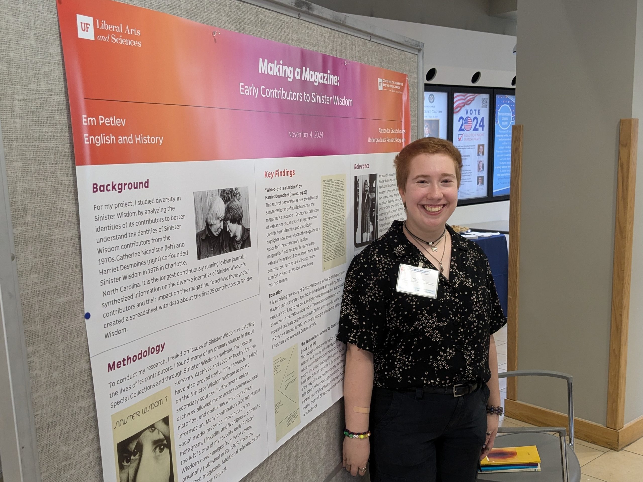 Em, dressed in a black floral buttoned shirt and black slacks, smiles in front of their presentation poster.