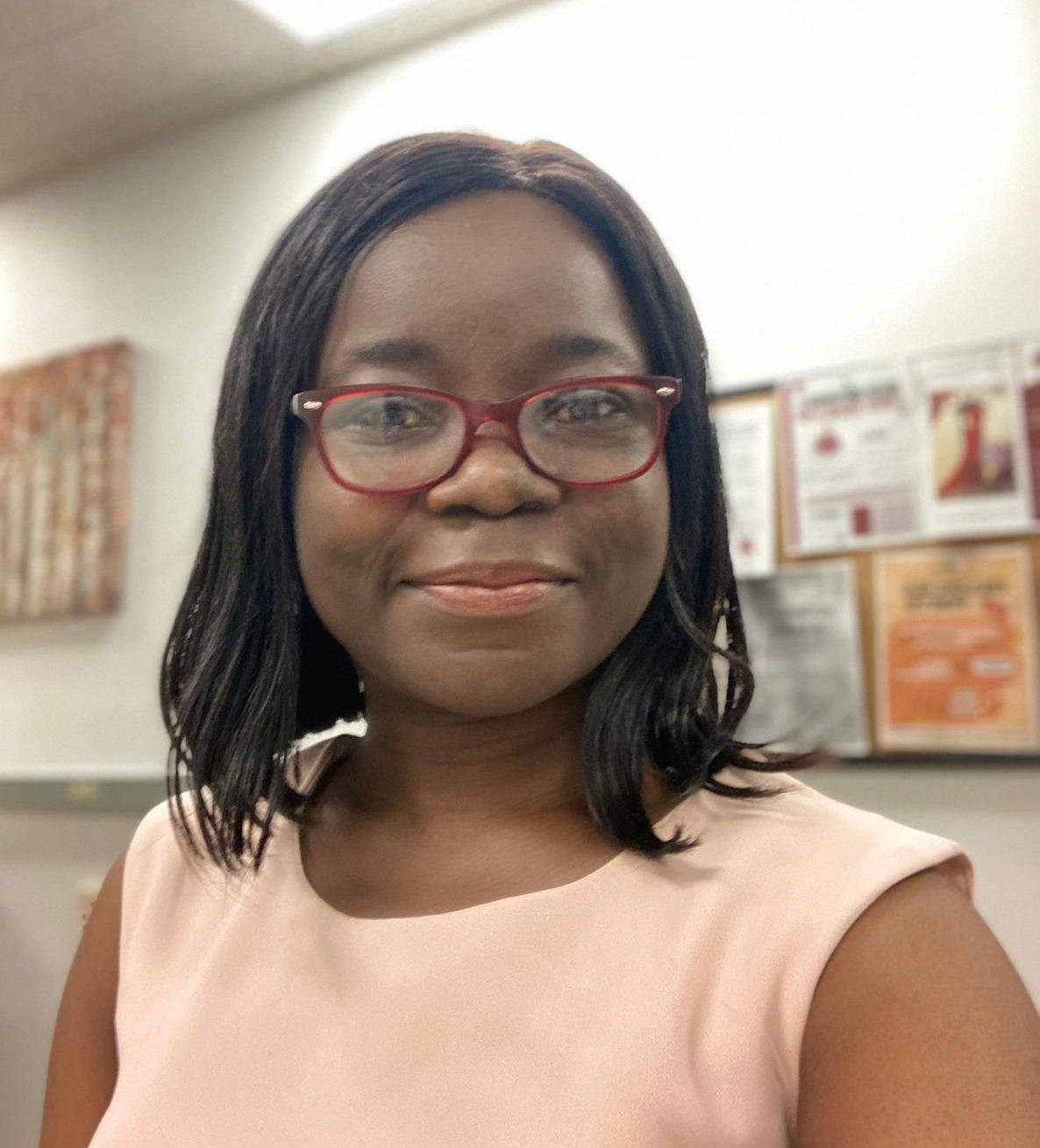 A headshot of smiling Abiola Emmanuel. She has dark skin and shoulder-length dark hair. She is wearing a cream colored sleeveless blouse and glasses with red frames.