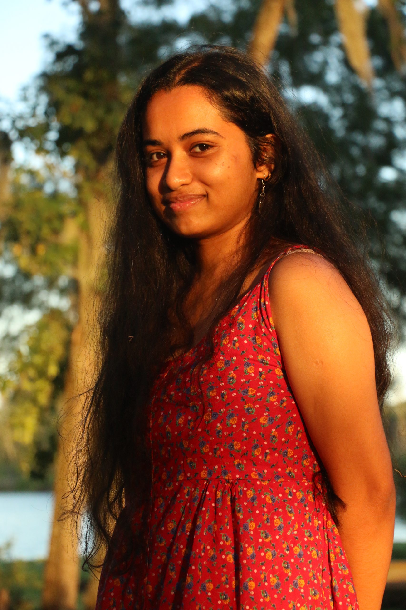Annesha Palit stands outside in the natural light. She has brown skin and long, dark hair. She wears a red, floral dress and earrings. Her arms are relaxed behind her back and she smiles at the camera.