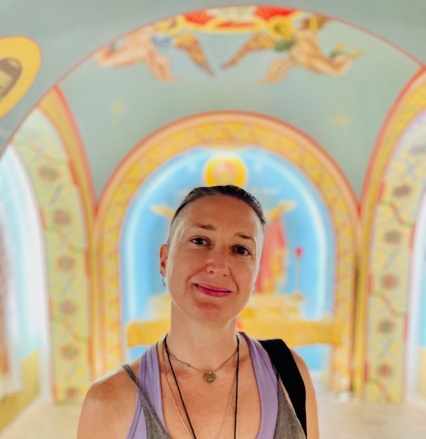 Daun Fields smiles in front of pastel frescoes painted on the surrounding walls and ceiling. She is a white woman with a brown undercut. She is wearing layered necklaces over her outfit.