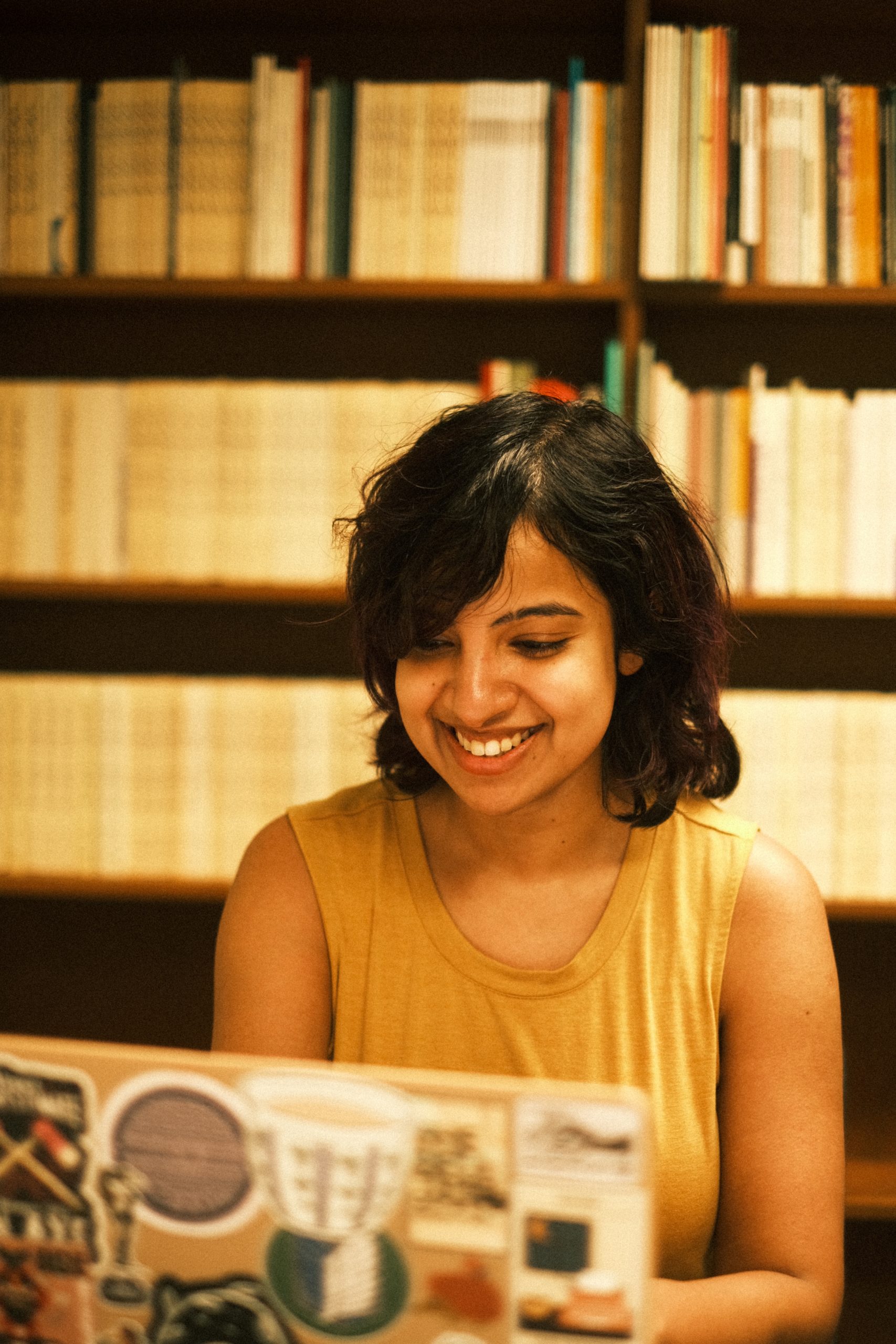 A candid photo of Debakanya Haldar smiling openly as she looks at her laptop. She is a South Asian woman with brown skin and dark shoulder-length hair. Her laptop is covered in stickers, and behind her are rows of books on a library bookshelf.