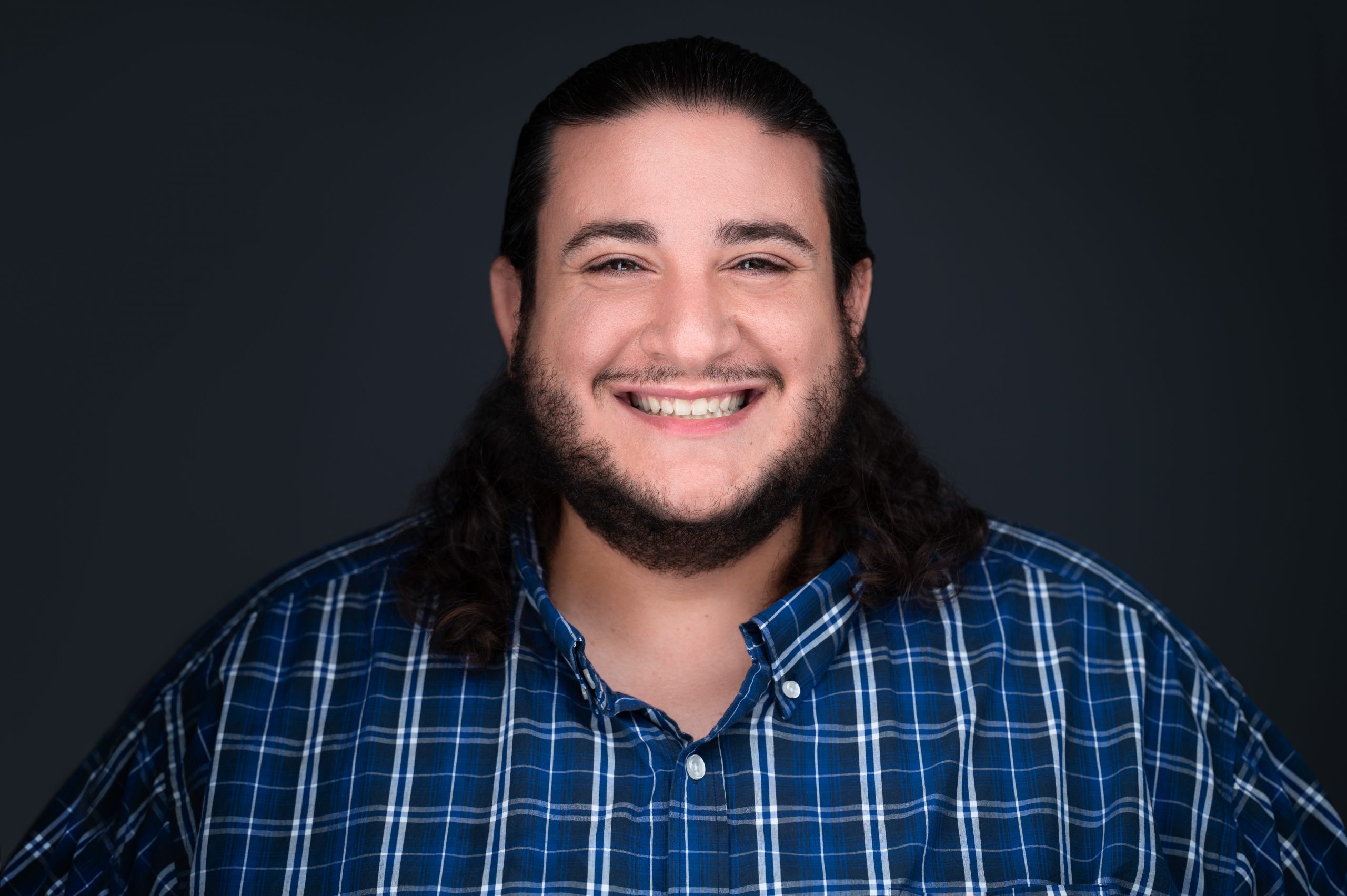A headshot of Jacob Domosh, who is a large, long-haired, tan man. He is wearing a blue and grey plaid shirt and smiling in front of a dark blue background
