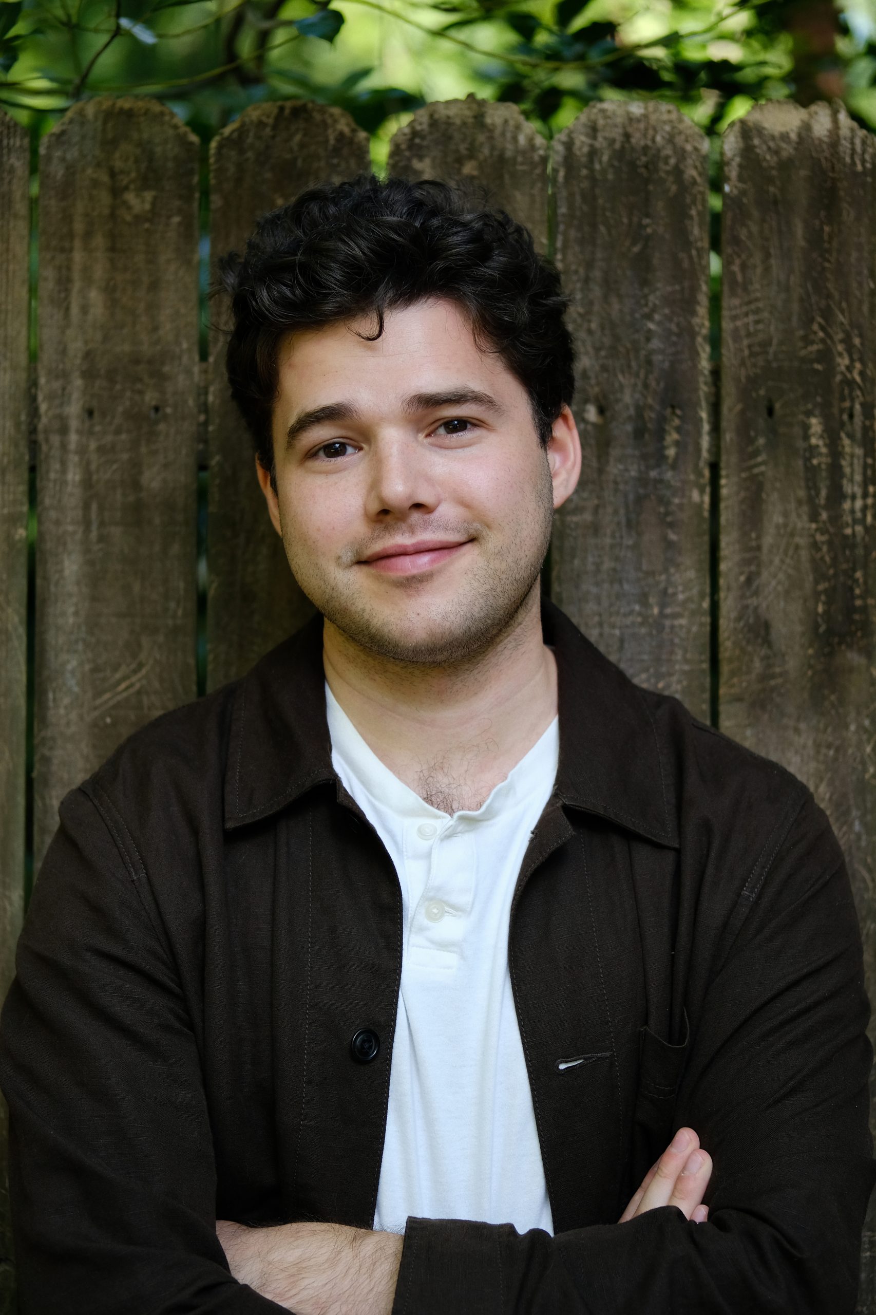 A headshot of Jonathan Wolf, who is wearing a white shirt under a dark, unbuttoned collared coat. He has dark and curly hair, light skin, a friendly smile, and crossed arms. He is standing in front of a wooden fence.