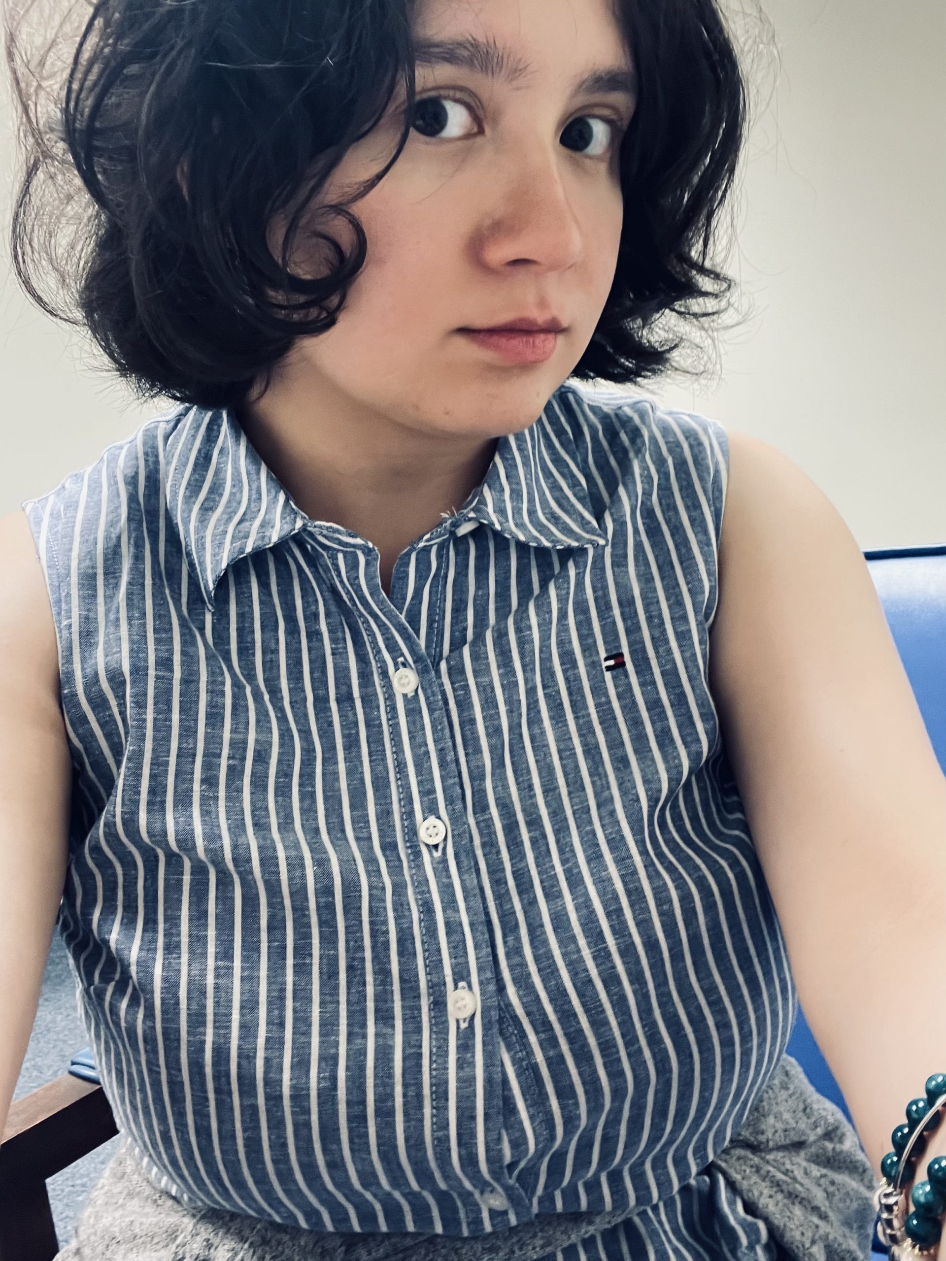 Kara looks at the camera. She has dark hair cropped just above her shoulders, and she is wearing a blue and white-striped dress.