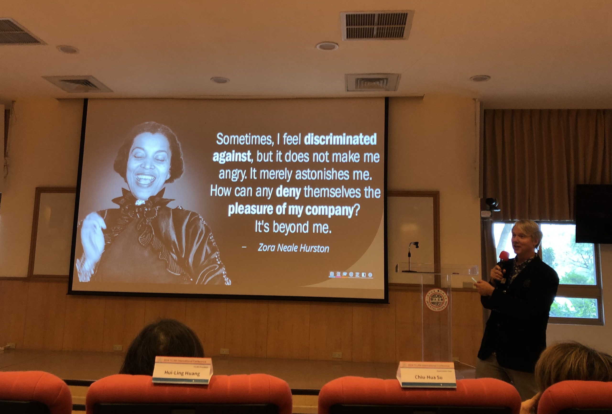 Kenneth Kidd, dressed in a dark blazer and light pants, stands in front of a projected slide, which features a picture of Zora Neale Hurston and the quote: "Sometimes, I feel discriminated against, but it does not make me angry. It merely astonishes me. How can any deny themselves the pleasure of my company? It's beyond me." Kidd is holding a microphone, mid-speech,