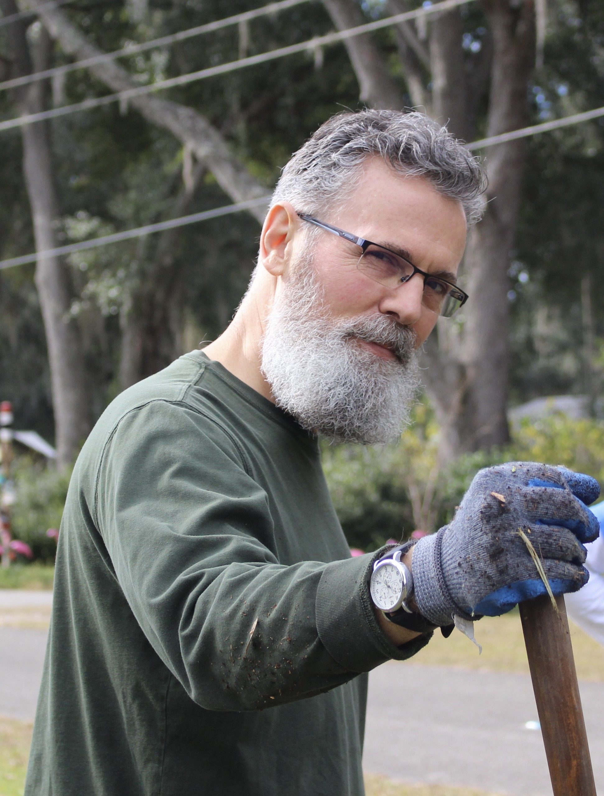 Terry Harpold smiles, holding the wooden handle of a gardening tool while planting trees. He wears a green sweater, silver wristwatch, blue gloves, and black rimless glasses.